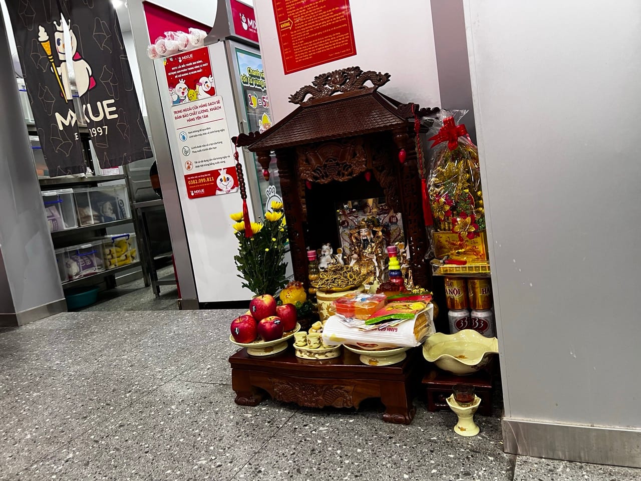 A traditional Vietnamese spirit altar (bàn thờ thần tài) inside a Mixue ice cream shop in Lao Cai — with fruit offerings, yellow flowers, and Hanoi beer cans