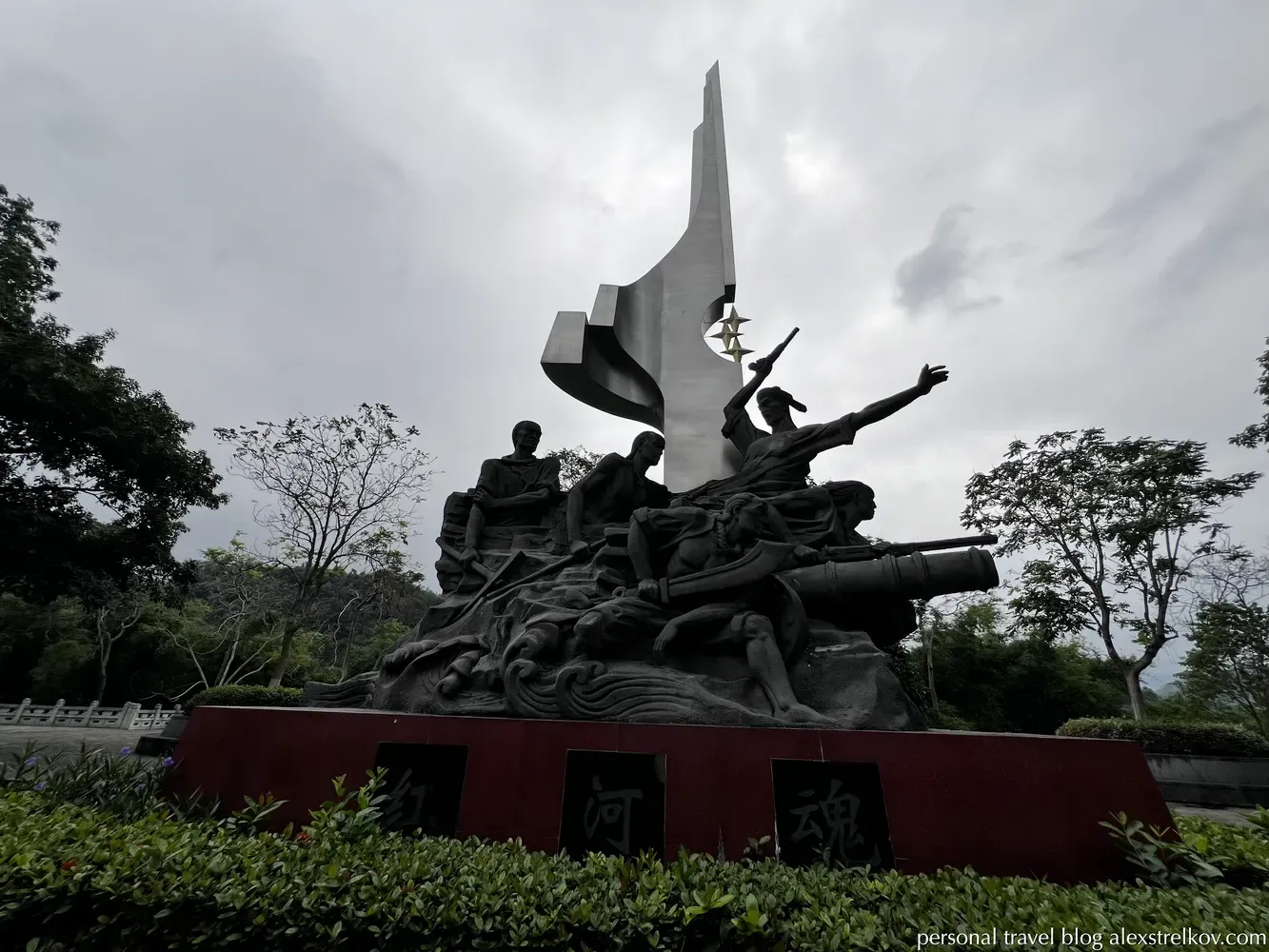 A dramatic bronze monument in Hekou, China, showing armed revolutionaries from 1908 with a cannon, set against a modern silver stele and cloudy sky.