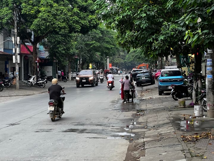 A tree-lined street in Lao Cai, Vietnam — motorcycles, cars, and locals going about their morning, with a blue VinFast EV parked on the right